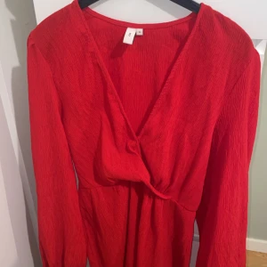 Röd omlottklänning  - Röd kort omlottklänning med lång ärm och v-ringad hals. Klänningen har en lätt rynkad struktur i tyget och markerad midja. Perfekt för dig som vill sticka ut med en färgstark look.