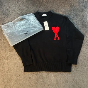 Svart stickad tröja från Ami Paris - Svart stickad tröja från Ami Paris med röd hjärtformad A-logga på bröstet. Tröjan har rund hals, ribbade muddar och är långärmad. Perfekt för dig som gillar stilrena plagg med en twist. Materialet är mjukt och håller värmen under kyliga dagar.