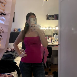 Rosa bandeau tubtopp - Säljer en snygg rosa bandeau tubtopp med rynkade detaljer framtill. Toppen är axelbandslös och har en tight passform som sitter skönt mot kroppen. Perfekt för dig som gillar färgstarka plagg och vill ha en enkel men trendig look.