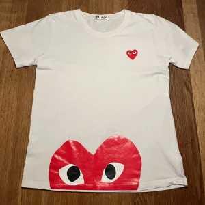 Vit t-shirt från Comme des Garçons - Säljer en vit t-shirt från Comme des Garçons med det ikoniska röda hjärtat med ögon på bröstet och en större version längst ner. Perfekt för en stilren och trendig look. Tar byten