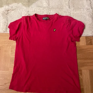 Röd t-shirt från Lyle & Scott Junior - Röd t-shirt från Lyle & Scott Junior med klassisk rund hals och korta ärmar. T-shirten har den ikoniska gula örnloggan broderad på bröstet. Tillverkad i mjuk bomull för en skön känsla. Perfekt för en avslappnad och stilren look.