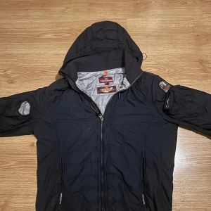 Parajumpers jacka - Mörkblå vindjacka från Parajumpers med huva och coolt PJS-märke på ärmen. Jackan har dragkedja framtill, två sidofickor med dragkedja och lätt fodrad insida i grått. Perfekt för dig som gillar stilren och funktionell design.