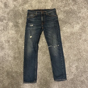 Mörkblå jeans från Nudie Jeans Co - Snygga mörkblå jeans från Nudie Jeans Co med slitna detaljer och klassiska orange sömmar. Modellen har raka ben, fem fickor och coola broderade detaljer på bakfickorna. Perfekt för dig som gillar en avslappnad streetstil.