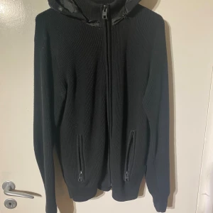 Svart stickad hoodie med dragkedja - Svart stickad hoodie från Jack & Jones med hel dragkedja framtill och två fickor med dragkedja. Huva i quiltad material ger extra stilpoäng. Perfekt för dig som gillar en clean look och vill ha något som är både bekvämt och snyggt.
