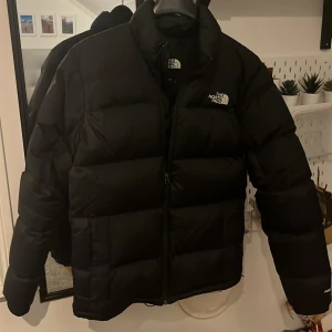 Svart dunjacka från The North Face - Säljer en svart dunjacka från The North Face i storlek S. Jackan har en klassisk pufferstil med hög krage, diskret logga på bröstet och ryggen samt dragkedja framtill. Perfekt för kalla dagar och riktigt snygg till streetwear-looken.