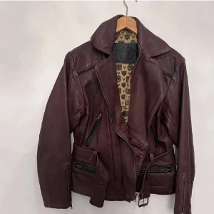 Mörkbrun skinnjacka med bälte - Snygg mörkbrun skinnjacka med bred krage, dragkedjor och bälte i midjan. Jackan har flera fickor med dragkedja och coolt mönstrat innerfoder. Perfekt för dig som vill ha en edgy och tidlös look.