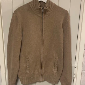 Beige stickad kofta från Dressmann - Säljer en beige stickad kofta från Dressmann i storlek M. Koftan har ribbad krage, muddar och fickor samt dragkedja framtill. Materialet är 79% bomull och 21% polyamid, vilket gör den mjuk och skön. Perfekt för lager-på-lager under kyliga dagar.