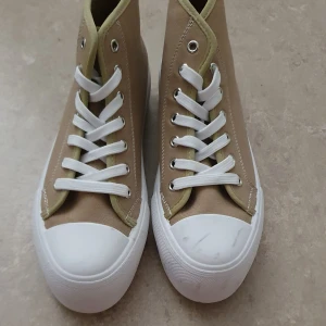Sneakers - Snygga beige sneakers med högt skaft och vit platt sula. Skorna har vita snören, silverfärgade öljetter och förstärkt vit tå. Tillverkade i canvasmaterial med ljusbeige kant runt öppningen. Perfekt för dig som gillar klassisk streetstil.