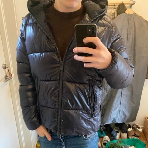 Blå pufferjacka från Save The Duck - Säljer en blå pufferjacka från Save The Duck i storlek S. Jackan är 100% animal free, har huva, dragkedja framtill och två fickor med dragkedja. Materialet är glansigt och syntetiskt, perfekt för kalla dagar. Snygg och modern look med quiltad design.