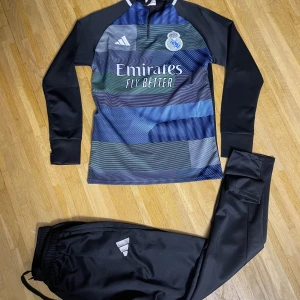 Real Madrid långärmad träningströja Adidas - Snygg långärmad Real Madrid träningströja från Adidas med grafiskt mönster i blått, grönt och svart. Tröjan har klubbmärke och Emirates Fly Better-tryck på bröstet. Materialet är slätt och sportigt, perfekt för fotbollsträning. Matchande svarta träningsbyxor med Adidas-logga ingår.