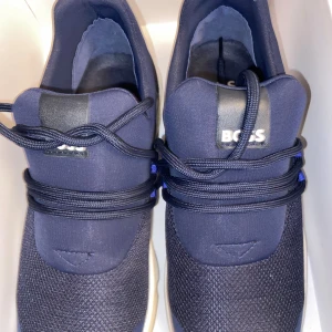 Marinblå sneakers från BOSS - Snygga marinblå sneakers från BOSS med vit sula och svart detalj på hälen. Ovandelen är i mesh och syntet, med rund tå och snörning. BOSS-loggan syns tydligt på hälen och plösen. Perfekt för dig som gillar stilrena och sportiga skor.
