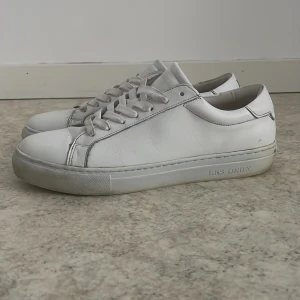 Vita sneakers från Les Deux i skinn - Snygga vita sneakers från Les Deux med rund tå och platt sula. Skorna är tillverkade i skinn och har snörning framtill. På hälen finns broderade detaljer i blått, vitt och rött. 