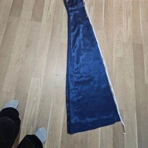 Marinblå långklänning med spetsdetaljer - Snygg marinblå långklänning i glansigt satinmaterial med hög hals och öppet nyckelhål framtill. Överdelen har vita spetsbroderier och pärldetaljer som ger en lyxig känsla. Klänningen har ärmlös design och markerad midja för en elegant siluett.