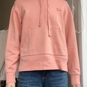 Rosa hoodie från Acne Studios - Säljer en rosa hoodie från Acne Studios med det klassiska face-logotypen på bröstet. Tröjan har huva med snörning, stor magficka och är tillverkad i mjuk bomull. Passformen är normal och ärmarna är långa.