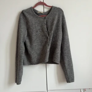 Grå stickad kofta från Gina Tricot - Mysig grå stickad kofta från Gina Tricot i storlek L. Koftan har rund hals, långa ärmar och knäppning med fyra knappar framtill. Materialet är mjukt och fluffigt, perfekt för kyliga dagar. Enkel och stilren design som funkar till många outfits.