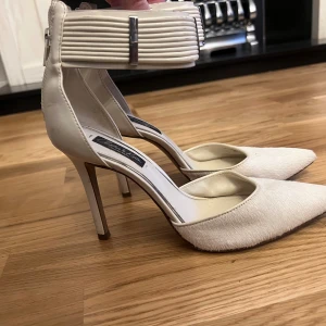 Beige pumps med spetsig tå och dragkedja - Snygga pumps från Charles & Keith i beige med spetsig tå och hög klack. Skorna har en bred, ribbad rem runt ankeln med metallspänne och dragkedja baktill. Materialet är en mix av skinn och mocka med en mjuk finish framtill.