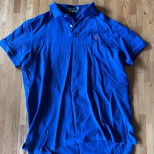 Blå pikétröja från Polo Ralph Lauren - Säljer en klassisk blå pikétröja från Polo Ralph Lauren med orange broderad logga på bröstet. Tröjan har korta ärmar, krage och knappar framtill. Tillverkad i bomull och passar perfekt till en avslappnad stil.