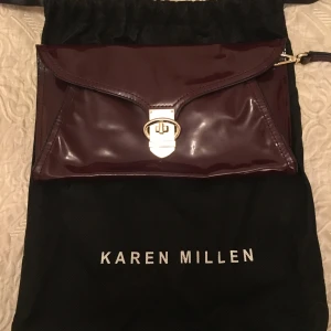 Vinröd kuvertväska från Karen Millen - Snygg vinröd kuvertväska i glansigt material från Karen Millen. Väskan har ett elegant guldigt spänne framtill och stilren design. Perfekt att bära i handen eller med den lilla remmen. En riktig statement piece för din outfit.