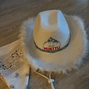 Vit Minttu cowboyhatt med glitter - Säljer en vit cowboyhatt från Minttu med glittrig yta och blått paljettband runt hatten. Hatten har fluffiga kanter och ett Minttu-märke framtill. Perfekt för temafest eller när du vill sticka ut med en unik accessoar.
