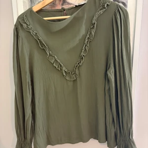 Olivgrön blus med volangdetalj - Säljer en olivgrön blus från Vero Moda med långärmade puffärmar och volangdetalj i V-form framtill. Blusen har rund halsringning och knapp i nacken. Materialet är mjukt och lätt polyester, perfekt för dig som gillar stilrena detaljer.