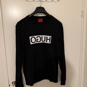 Svart långärmad tröja från Hugo - Svart långärmad tröja från Hugo med vit logga tryckt i spegelvänd stil på bröstet. Tröjan har en klassisk rund halsringning och är tillverkad i mjuk bomull, perfekt för en clean och stilren look.