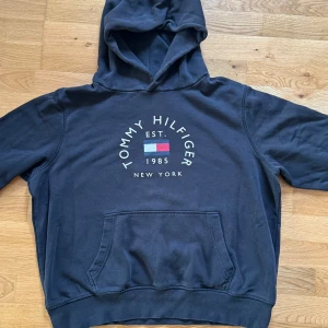Mörkblå hoodie från Tommy Hilfiger - Snygg mörkblå hoodie från Tommy Hilfiger med klassisk logga och text framtill. Tröjan har huva, magficka och är tillverkad i en mjuk ekologisk bomullsblandning. Perfekt för chill dagar och streetstyle.