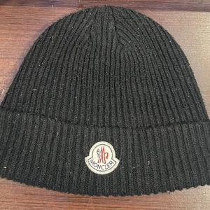 Svart ribbstickad mössa från Moncler - Svart ribbstickad mössa från Moncler med klassisk Moncler-logga framtill. Mössan är tillverkad i ett mjukt och värmande material och har en uppvikt kant för extra stilpoäng. Perfekt accessoar för kyliga dagar.