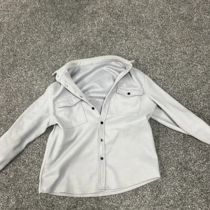Ljusgrå overshirt med knappar - Snygg ljusgrå overshirt med svarta knappar och två bröstfickor med lock. Skjortan har klassisk krage och långa ärmar med knapp vid ärmslut. Perfekt att slänga över en t-shirt för en chill look.