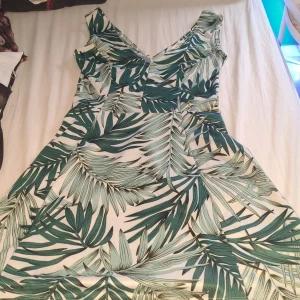 Grönmönstrad sommarklänning  - Snygg kort klänning från H&M i storlek S med tropiskt bladmönster i grönt och vitt. Klänningen har v-ringad hals och är ärmlös, perfekt för varma dagar. Tillverkad i mjuk bomull som känns skön mot huden och har en lätt, flowy passform.
