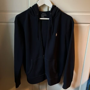 Mörkblå hoodie Polo Ralph Lauren - Snygg svart hoodie från Polo Ralph Lauren med dragkedja och klassisk logga på bröstet. Tröjan har huva med snörning och två fickor framtill. Storlek S och den är i väldigt bra skick!