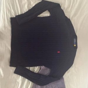 Mörkblå stickad tröja Polo Ralph Lauren - Säljer en mörkblå kabelstickad tröja från Polo Ralph Lauren med klassisk röd broderad logga på bröstet. Tröjan har rund halsringning, långa ärmar och är tillverkad i mjuk bomull. Perfekt för dig som gillar stilren och tidlös design.