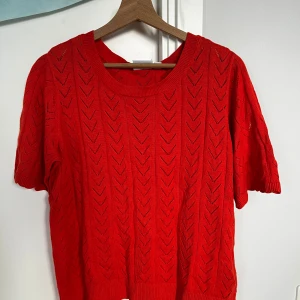 Röd stickad topp från VILA - Snygg röd stickad topp från VILA med kort ärm och rund halsringning. Tröjan har ett genomgående hålmönster i pilformat och ribbade kanter. Perfekt för dig som vill ha en färgstark och luftig look till våren eller sommaren.