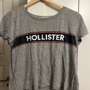 Grå Hollister t-shirt XS - Snygg grå t-shirt från Hollister i storlek XS. T-shirten har korta ärmar och ett stort tryck med Hollister-loggan över bröstet i vitt, blått och rött. Tillverkad i mjuk bomull och har en klassisk passform med rund hals.