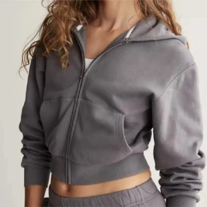 Grå croppad hoodie med dragkedja - Grå croppad hoodie med huva och dragkedja framtill. Tröjan har långa ärmar, ribbade muddar och en stor ficka på magen. Perfekt för en chill och avslappnad stil. Liten i storleken.