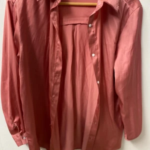 Rosa långärmad skjorta i satin - Snygg rosa skjorta i satinliknande material med klassisk krage och knäppning framtill. Skjortan har långa ärmar med manschett och en lösare passform, perfekt för en trendig och avslappnad look.