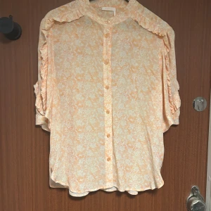 See by Chloe Blommig volangskjorta i rosa och orange - Blus från See by Chloé i ljusaprikos och vit med blommigt mönster. Skjortan har volangdetaljer längs axlarna, knappar framtill och korta ärmar. Luftigt material som känns lyxigt och flowy.