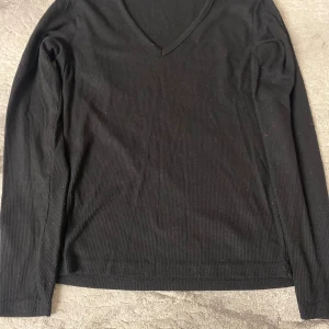 Svart ribbad långärmad tröja - En svart långärmad tröja med v-ringning och ribbad struktur. Perfekt basplagg med enkel och stilren design, passar till många olika outfits. Mjuk och skön att bära.