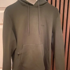 Olivgrön hoodie från BOSS - Säljer en stilren olivgrön hoodie från BOSS med broderad logga på bröstet. Tröjan har huva med dragsko, magficka med dragkedjedetalj och långa ärmar. Perfekt för en chill och avslappnad look.