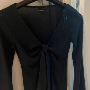 Mörkblå långärmad topp med knyt - Snygg svart långärmad topp från Gina Tricot med djup v-ringning och knyt framtill. Toppen har en slim passform och är perfekt för att skapa en trendig look. Tillverkad i mjuk och stretchig polyester för en bekväm känsla.