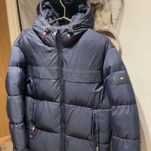 Mörkblå pufferjacka från Tommy Hilfiger - Snygg mörkblå pufferjacka från Tommy Hilfiger med broderad logga över bröstet och patch på ärmen. Jackan har huva med dragsko, två fickor med dragkedja och hel dragkedja framtill. Perfekt för kalla dagar och riktigt stilren look.             Nypris 3000kr