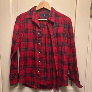 Ralph Lauren skjorta - Säljer en klassisk röd rutig skjorta från Ralph Lauren med blå broderad logga på bröstfickan. Skjortan har långa ärmar och knappar hela vägen Perfekt för dig som gillar en tidlös och avslappnad stil. Storlek L för barn så den passar dig mellan 160-170 beroende på vilken passform du gillar!😊