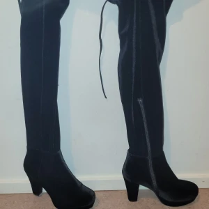 Svarta höga stövlar med klack. Från Skopunkten (Aldrig använda).  - Säljer ett par svarta, höga stövlar från Skopunkten i storlek 38. De har en snygg klack samt dragkedja på sidan. Materialet är glansig syntet som ger en elegant look. Stövlarna har snörning upptill för extra stilpoäng. Skorna är aldrig använda.