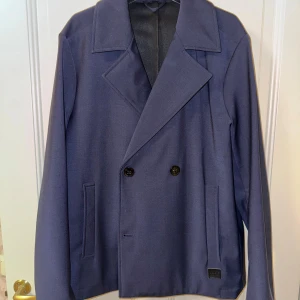 Mörkblå skepparkavaj från Dondup - Säljer en stilren mörkblå skepparkavaj från Dondup, Made in Italy. Jackan har klassisk dubbelknäppning, bred krage och raka fickor framtill. Materialet är mjukt och känns lyxigt, perfekt för dig som gillar minimalistisk och snygg design.
