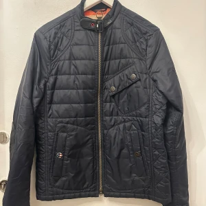 Svart quiltad Barbour jacka S - Quiltad svart jacka från Barbour Steve McQueen Collection med coola detaljer som axelrem med spänne, dragkedja och knappar. Jackan har fickor med lock, förstärkta armbågar i skinn och orange foder. Perfekt för dig som gillar klassisk stil med edge.