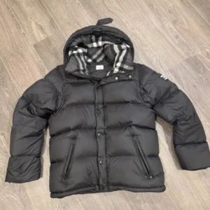 Svart Burberry pufferjacka med huva - Svart pufferjacka från Burberry med stor patch på ärmen och klassiskt rutigt foder i huvan. Jackan har dragkedja och knappar framtill, två fickor med dragkedja och en skön, vadderad känsla. Perfekt för kalla dagar och riktigt snygg streetstil.