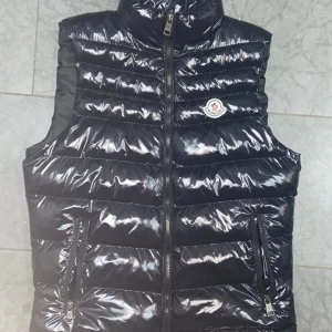 Svart dunväst från Moncler - Snygg svart dunväst från Moncler med glansig finish och klassisk logga på bröstet. Västen har två dragkedjefickor framtill och quiltad design som ger en cool och sportig look. Perfekt att slänga över en hoodie eller tröja när det är kyligt ute.