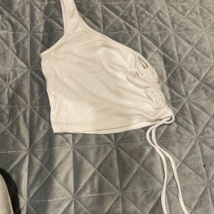 Vit oneshoulder croppad topp från FB Sister - Säljer en vit ribbad oneshoulder topp från FB Sister i storlek XS. Toppen är croppad med snörning på sidan för att justera passformen. Perfekt för dig som gillar trendiga och unika detaljer. Materialet är mjukt och stretchigt, vilket gör den superbekväm.