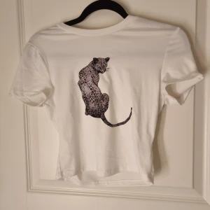 Vit t-shirt med leopardtryck - Snygg vit t-shirt med ett stort leopardtryck på bröstet. Modellen är kortärmad och har en klassisk rund hals. Materialet känns mjukt och skönt, perfekt för dig som gillar djurmotiv och vill sticka ut lite extra.