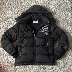 Svart pufferjacka från Mercier - Svart pufferjacka från Mercier med huva och coola patches på bröstet. Jackan har dragkedja framtill, två fickor med dragkedja och ribbade muddar vid ärmsluten. Perfekt för kalla dagar och riktigt snygg streetstil.