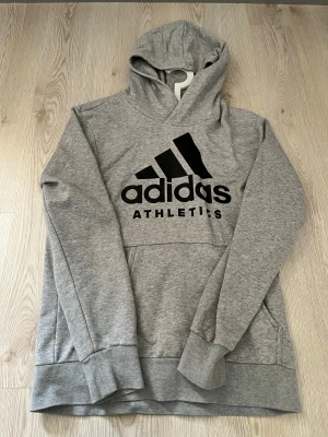 Grå Adidas hoodie med svart logga - Snygg grå hoodie från Adidas med stor svart logga och texten ATHLETICS på bröstet. Tröjan har huva, lång ärm och en praktisk magficka. Perfekt för chill eller sportiga dagar, riktigt skön och stilren design.
